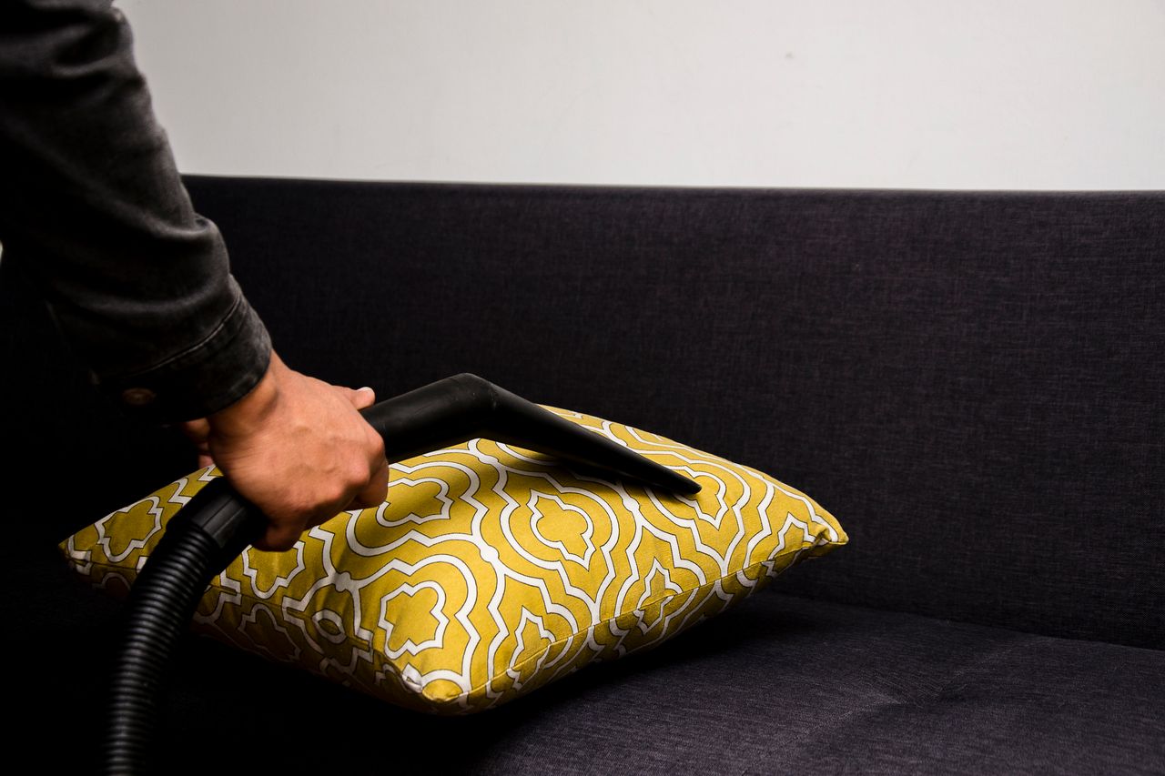 team-01 Close-up view of a person vacuuming a patterned pillow on a dark sofa indoors.